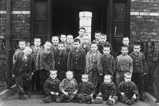 A black and white photo of about two dozen pre-adolescent boys in uniform jackets standing or sitting outside a brick building with what appear to be metal doors