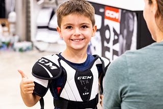 A young boy wearing hockey shoulder pads smiles and holds up a thumbs up. He is white with short brown hair. An adult woman with brown hair tied back in a ponytail and green shirt smiles at the boy with her back turned to the camera