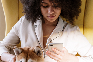 This woman in her pajamas looking at her phone has a dog by her side.