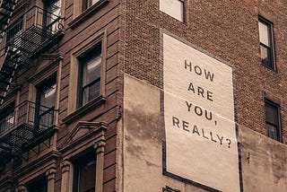 A tall brick building in a city with a sign that reads “how are you, really?”