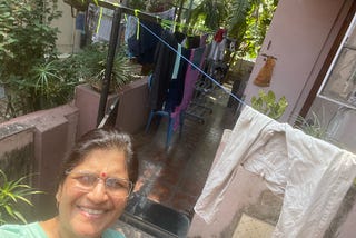 A woman wearing a red dot on her forehead (Southeast Asian Indian) smiles at the camera. There are clotheslines behind her where clothes dry. There are bright green plants beside the lady and behind the clotheslines. It is a bright, sunny day in India.