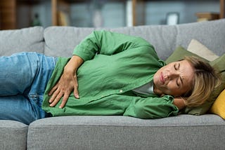 Mature woman alone at home sick, holding hands to her stomach, lying on a sofa.