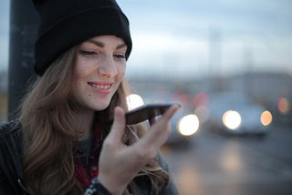 Girl with black hat on holding phone up and smiling with busy street of cars in the background