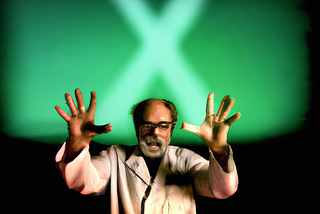 A surgeon in a bloodied white coat, stands with a green screen behind him, gestures with his hand as he addresses an unseen audience.