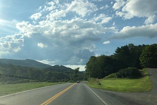 A highway in Vermont, lined with trees, and white clouds and mountains in the distance against a blue sky.