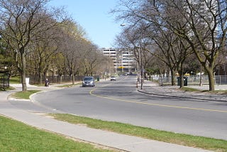 City street in daytime with a treelined streets and a car in the distance.