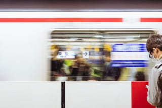 A man wearing a face mask looks at his phone while a train zooms by in the Tokyo subway.