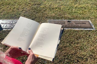 An open book is held in front of a gravestone. Long white hair and hands of the person reading are visible at the corner of the image.