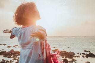 Woman with short hair & arms outstretched to the ocean, with the sun beaming on her face and upper body. Face to the sky.