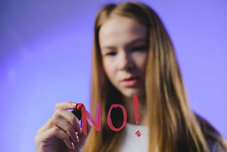 A girl writing no on the screen with a pink pen. She is lit with purple background.