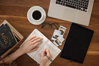 A person writes in an open journal, surrounded by coffee, glasses and a computer