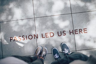 A black-and-white image from the point of view of people looking down shows their feet with the words “Passion led us here” in red as if engraved on a sidewalk.