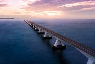 A two-lane highway stretching away over a body of water, toward a sunset in the distance.