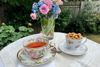 A floral teacup filled with tea, and a small demitasse cup filled with coconut granola on a small table in the garden. A bouquet of lavendar roses and blue hydrangeas is in the background.