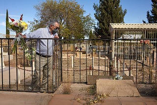 An 80 year old man holds a flower bouquet in his hand as he bends over a small fence to observe his father’s grave to which he hasn’t been to visit in almost 30 years. It is a cold and sunny day in winter at Cananea, Sonora’s cemetery. Most of the graves there are abandoned.