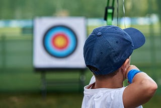 Boy wearing blue cap, blue armband, and white T-shirt aiming his bow and arrow at a target in the background.