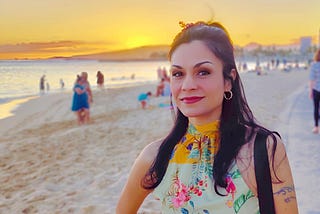 This image shows a person at a beach during sunset. They’re wearing a colorful floral-patterned top with yellow, pink, and green elements. The background features a beautiful golden-orange sunset over water, with other beachgoers visible in the distance. The beach has light-colored sand, and the atmosphere appears warm and peaceful. The person is smiling at the camera and has dark hair, hoop earrings, and appears to be enjoying the scenic beach environment.