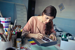 A woman with long dark hair wearing a peach shirt painting on a canvas at her desk.