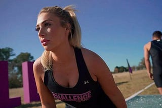 A focused female contestant from MTV’s ‘The Challenge’ is captured mid-competition, wearing a black sports tank with the show’s logo. She looks off into the distance with determination, highlighting her readiness for the upcoming challenge. In the background, a male contestant is seen, along with large purple letters that spell “OLIVIA,” possibly part of the show’s set design in an outdoor, sunny field.