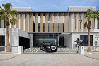 A cubist-inspired modern mansion with white walls, large square windows and large louvred panelling on the top floor. In the driveway sits a black Rolls Royce. To the right of the Rolls, there’s a graffitied statue of a gorilla standing on a plinth. Blue skies and tall palm trees indicate that this house is somewhere hot.