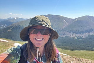 The author is in the mountains wearing a green hat, sunglasses, and a colorful tie-dye shirt with a backpack on his shoulders. He’s smiling.