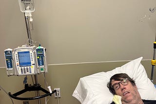 man in hospital bed with mouth open next to medical equipment.