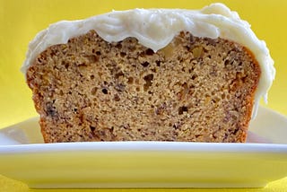 Image of banana bread on a white plate. a piece has been cut off from the end revealing the inside. the banana bread is frosted with a cream cheese frosting. it is sitting on a white plate. the background is bright yellow.