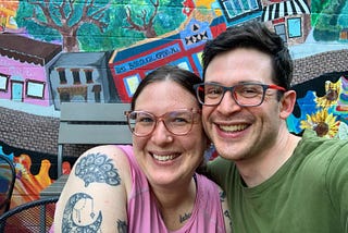 A photo of myself (a smiling white woman with pink glasses, brown hair, and tattoos) and my husband (a smiling white man with brown hair and glasses wearing a green shirt. We are sitting in front of a colorful mural of houses and trees