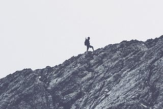 Silhouette of a mountaineer standing on an ascending ridge; photo all black & white.