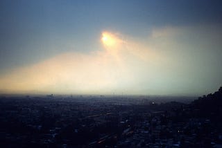 The valley of Mexico City with a dark cloud above and the sun partially shining opaquely through the clouds.