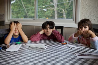 Three children sit at a table, one with hands over their eyes, another covering their ears, and the third with hands over their mouth.