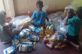 Three children sit on a floor with their backs to the camera surrounded by food goods from a humanitarian food basket.