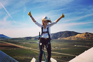 A blonde woman stands on top of a wind turbine with her arms raised