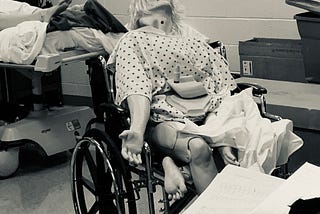 This is a sepia-toned photo of a nursing school manikin, that was taken in a medical learning facility/lab. The manikin has long shaggy hair, a hospital gown, and is sprawled out in a wheelchair in a very uncomfortable and unnatural looking position. The mood seems ominous.