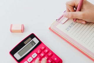 picture of hand writing on pad next to pink calculator showing 39 and pink eraser lying next to it