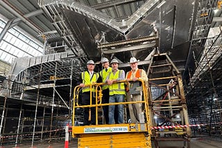 Four men stand on a lift underneath the hull of a large ferry under construction in a shipyard.