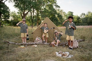 Group of children dressed as scouts at a campsite. They are surrounded by camping gear with a tent set up behind them. One child is playing the ukulele, another is reading a book, while the others are saluting. They are outdoors in a grassy area with trees in the background.