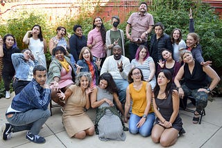 Photograph of 22 members of the HIP staff, outside in the afternoon during our 2022 retreat. Staff are assembled in 3 rows, with those in front kneeling on the pavement, the middle row seated in folding chairs, and the back row standing. Staff are making various “silly” poses/faces (sticking out tongues, crossing eyes, scrunching their noses, etc)