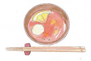 A bowl of reddish soup with a quartered lemon floating in it. A pair of wooden chopsticks are on a chopstick rest next to it.