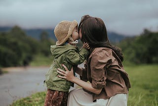 The image depicts a tender moment between a mother and her young child in a serene outdoor setting. The child, wearing a straw hat and a light green jacket, stands facing the mother and places a small kiss on her lips. The mother, dressed in a brown jacket and white blouse, holds the child gently, her expression unseen as she faces away from the camera. They are surrounded by a lush, green landscape under a cloudy sky, enhancing the intimate and peaceful atmosphere of the scene.