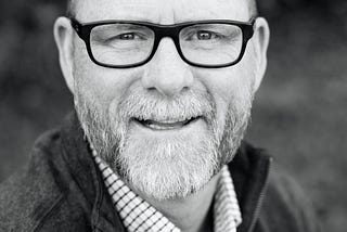 Black and white picture of an older man who is bald and wearing glasses and has a beard with gray hair.