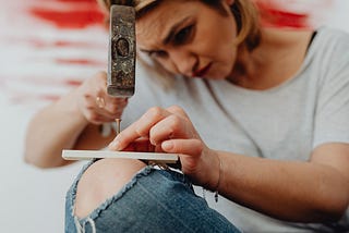 A woman about to try hitting a wood screw with a hammer into a thin piece of wood balanced on top of her leg