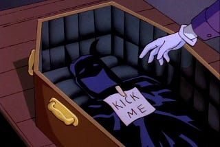 A screenshot from ‘Batman: The Animated Series’. The Joker’s hand can be seen attaching a “kick me” sign to Batman’s cape and cowl, inside a coffin.