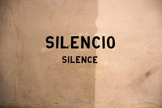 An off-white, roughly plastered wall, with the words ‘silencio’ and ‘silence’ stencilled onto it in black paint.