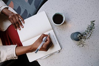 Black hands writing with a black pen on a blank page of a lined notebook. Sitting at a white table with a coffee and a sprig of rosemary in a little vase on the table.