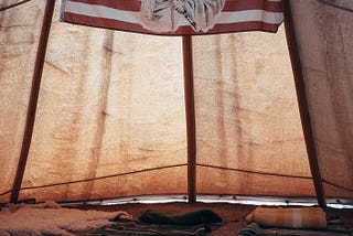 An image of a Native American elder imposed in the middle of an United States flag hangs from the inside of a tent styled in the tradition of an Indigenous tribe, complete with fur pelts, blankets, and a stone bordered fire pit on the earth beneath.