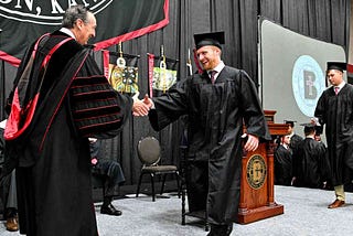 Benedictine College President Stephen D. Minnis on a stage shakes hands with a 2024 male graduate of the Catholic college in Atchison, KS.