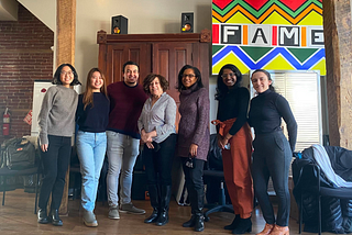 Seven people from the CMU Design Team and FAME staff stand together, smiling, in a spacious, cozy office room decorated with a colorful painting of the FAME logo.