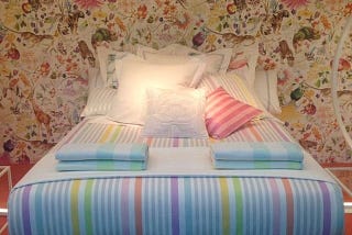 A picture of the Zara Home shop, with a bed with white bedlinen, with colorful stripes (purple, blue, grey, yellow, green, pink and orange). Lots of white cushions complete the bed.