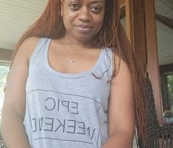 A young African American woman with long, braided hair and large, expressive eyes smiles at the camera. She is inside a house, wearing a sleeveless T-shirt that says “EPIC WEEKEND,” with the image appearing in reverse in the image.